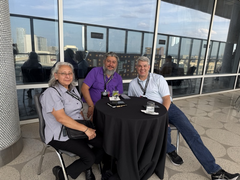 Three guests at a table on the outdoor patio during the welcome reception, Dallas high-rises behind them